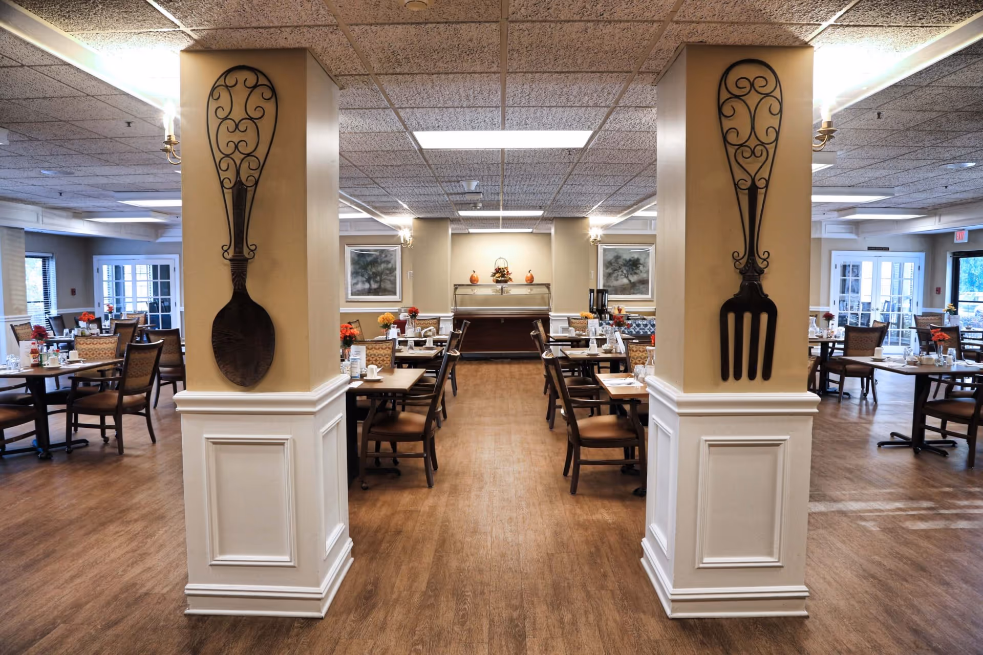 A spacious dining room in a senior living facility with wooden floors, multiple tables and chairs arranged neatly, and decorative large spoon and fork wall hangings on two central pillars. The room is well-lit with ceiling lights and has floral centerpieces on the tables.