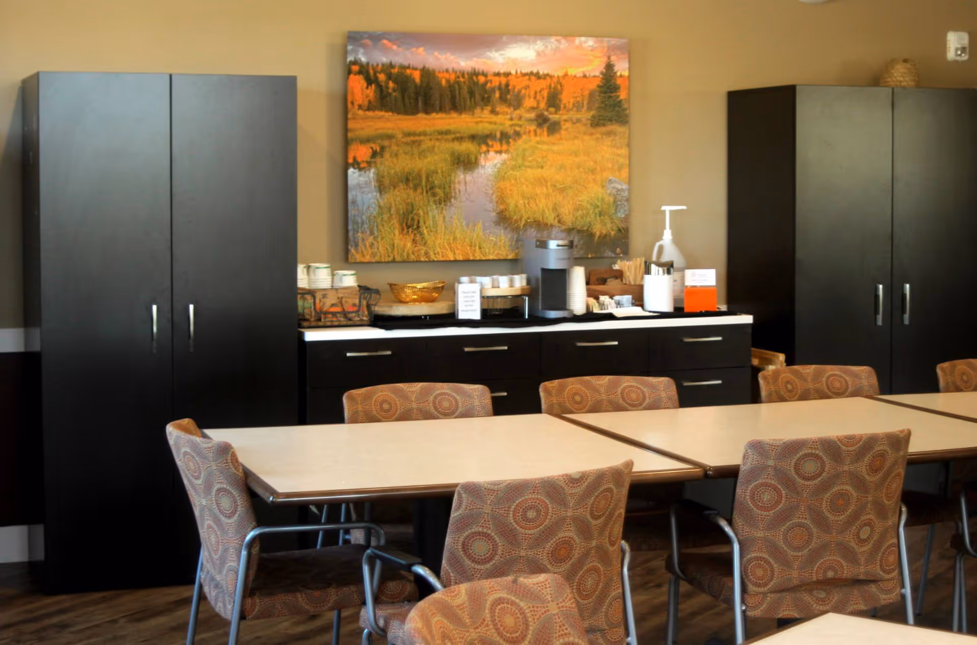 A dining area with several tables and patterned chairs arranged around them. Against the wall are two tall black cabinets and a black sideboard with cups, a coffee machine, and other beverage supplies. Above the sideboard hangs a large landscape painting depicting a serene autumn scene with trees and a river.
