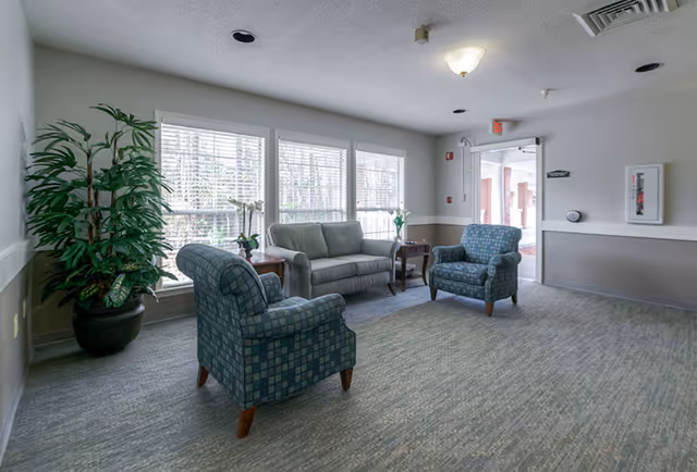 A bright sitting area with a large window letting in natural light. The room features a gray sofa, two patterned armchairs, two small wooden side tables with plants and flowers, a large potted plant in the corner, and a carpeted floor. The walls are painted light gray with a white trim, and there is a ceiling light fixture. An open doorway leads to another area of the facility.