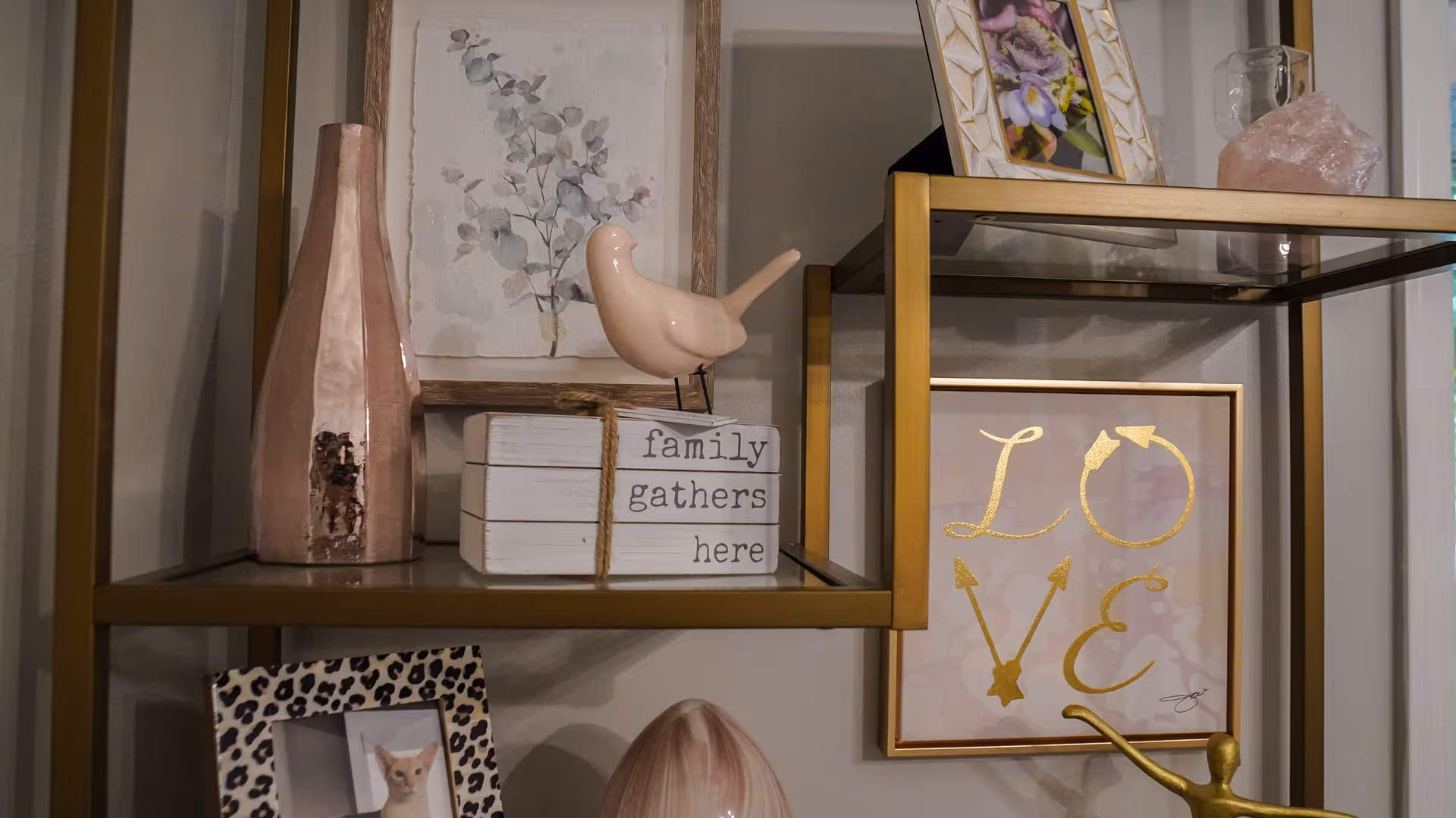 Decorative shelving unit with various home decor items including a pink ceramic bird, a pink vase, a framed floral print, a framed picture with the word LOVE in gold letters, a stack of wooden blocks with the words 'family gathers here', and a photo frame with a picture of a small dog.
