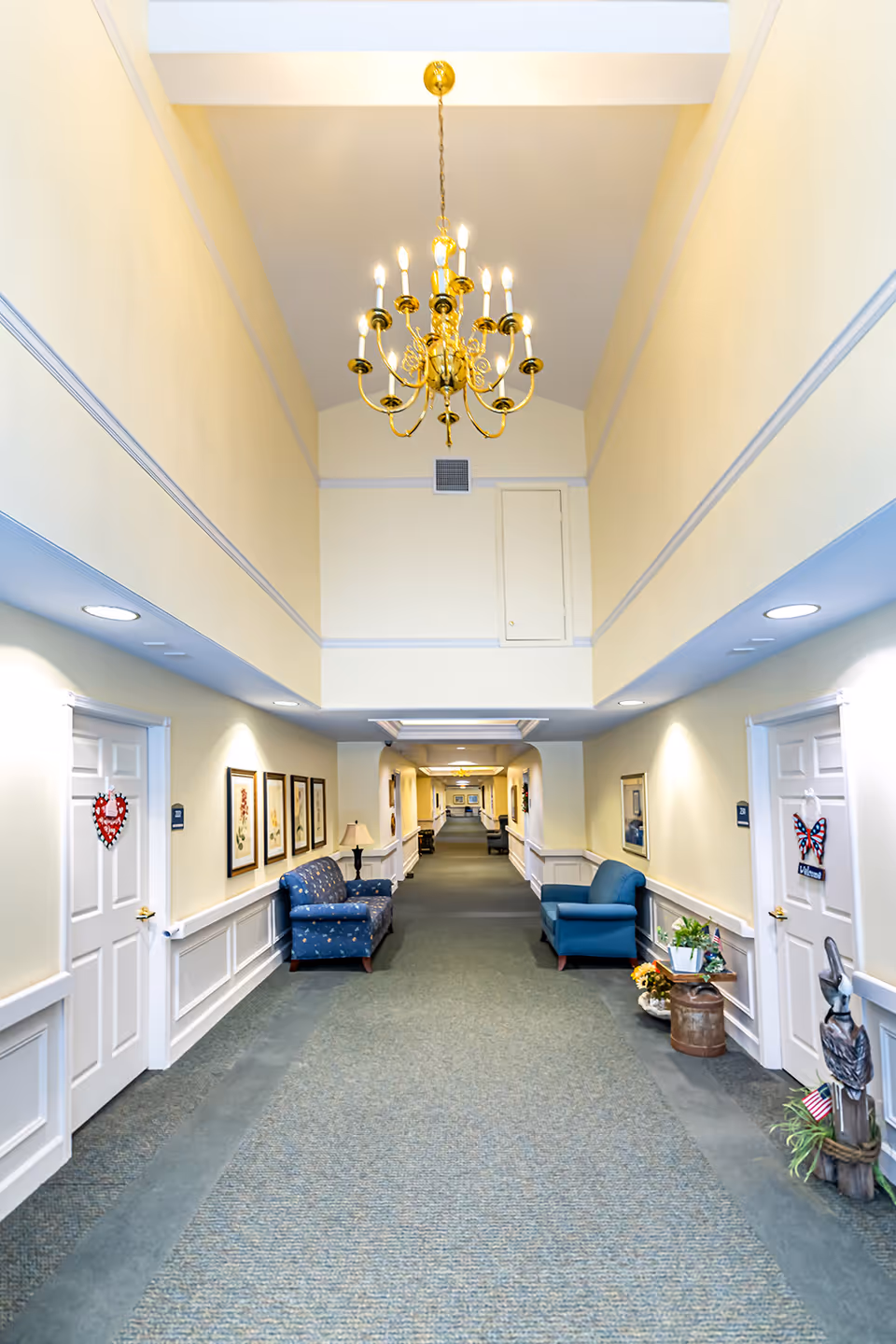 A long, well-lit hallway in an assisted living facility with a high ceiling and a gold chandelier. The hallway has light yellow walls with white trim, carpeted floors, and several framed pictures on the walls. There are two blue upholstered chairs and a small table with a lamp on the left side, and another blue chair with a small plant and decorative items on the right side. Doors with decorative wreaths are visible on both sides of the hallway.