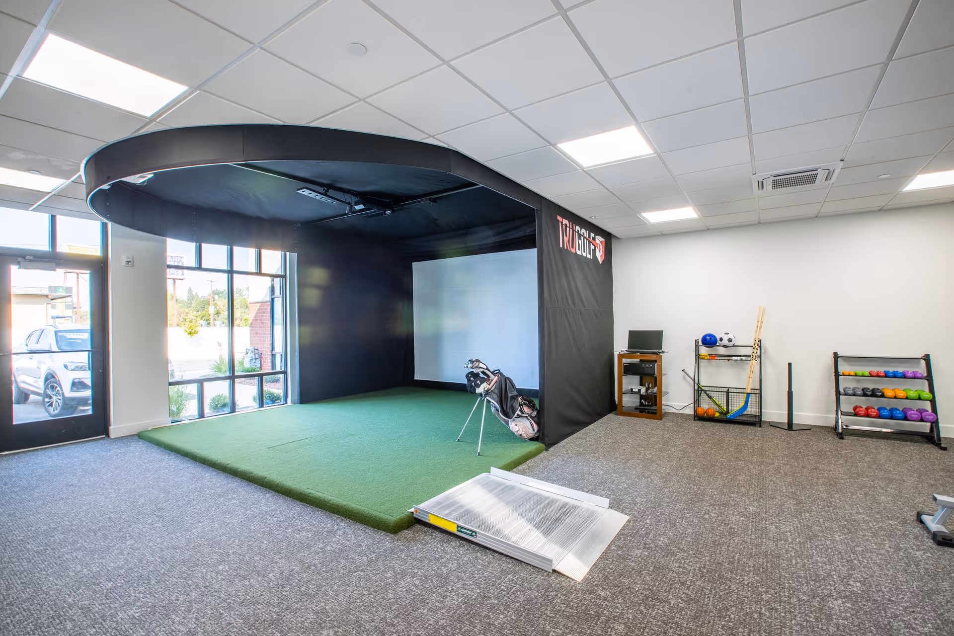 Indoor room with a golf simulator setup featuring a green artificial turf platform and a large screen inside a black enclosure labeled 'TRUGOLF'. Nearby, there is a rack with various sports balls and equipment, a small wooden cabinet with a computer monitor, and a ramp leading onto the turf area. Large windows and a glass door allow natural light into the space.