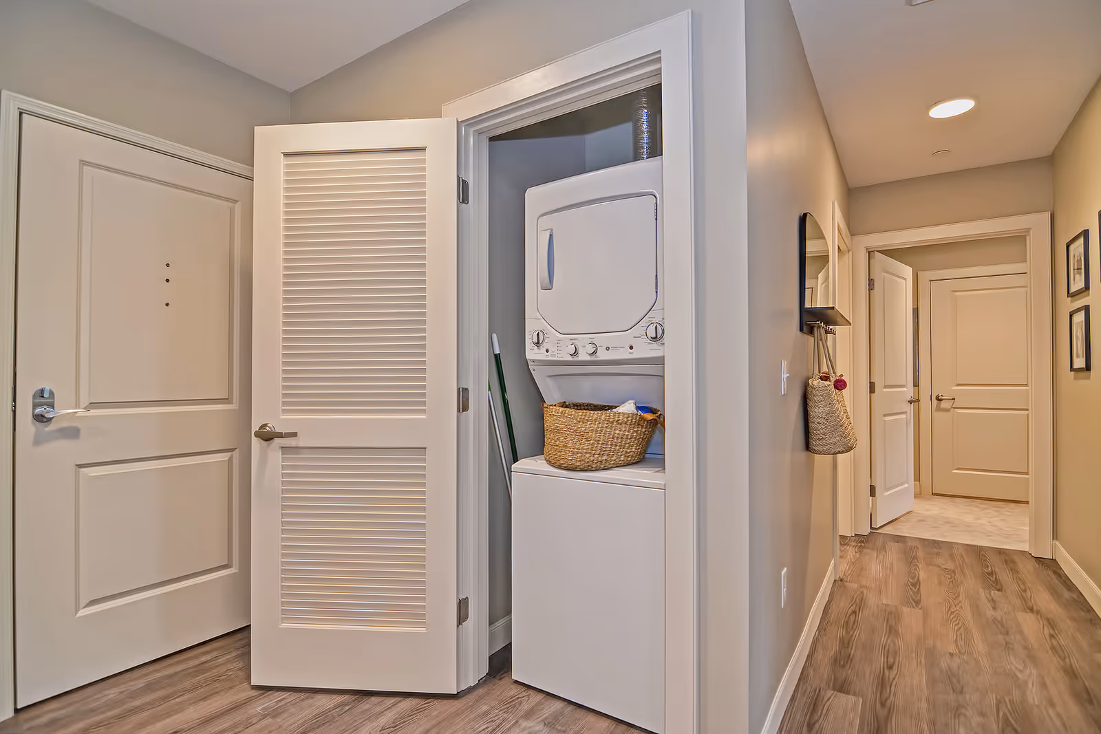 A hallway in a retirement community with light beige walls and wood flooring. On the left side, there is a closet with louvered doors open to reveal a stacked washer and dryer unit with a wicker laundry basket on top. The hallway extends to the right with several closed doors and a wall-mounted mirror with a hanging woven bag underneath.