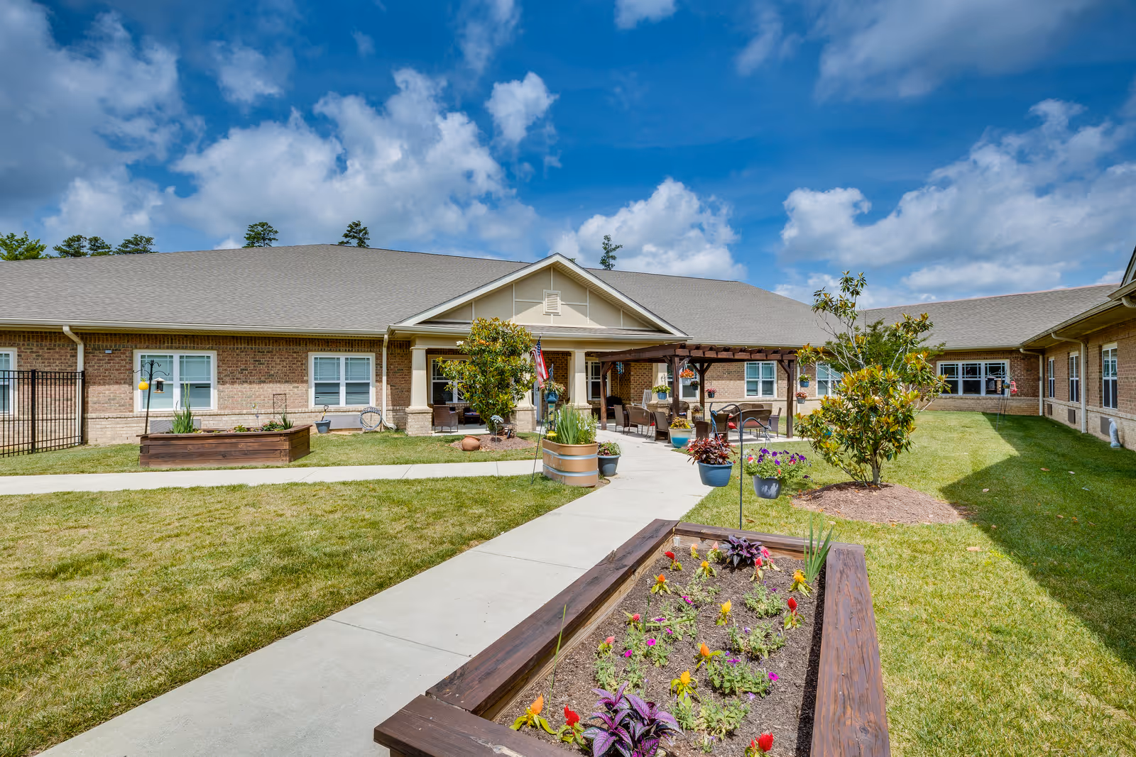 Single-story brick assisted living building with a courtyard, raised flower beds, a walkway, and a covered entrance under a blue sky.