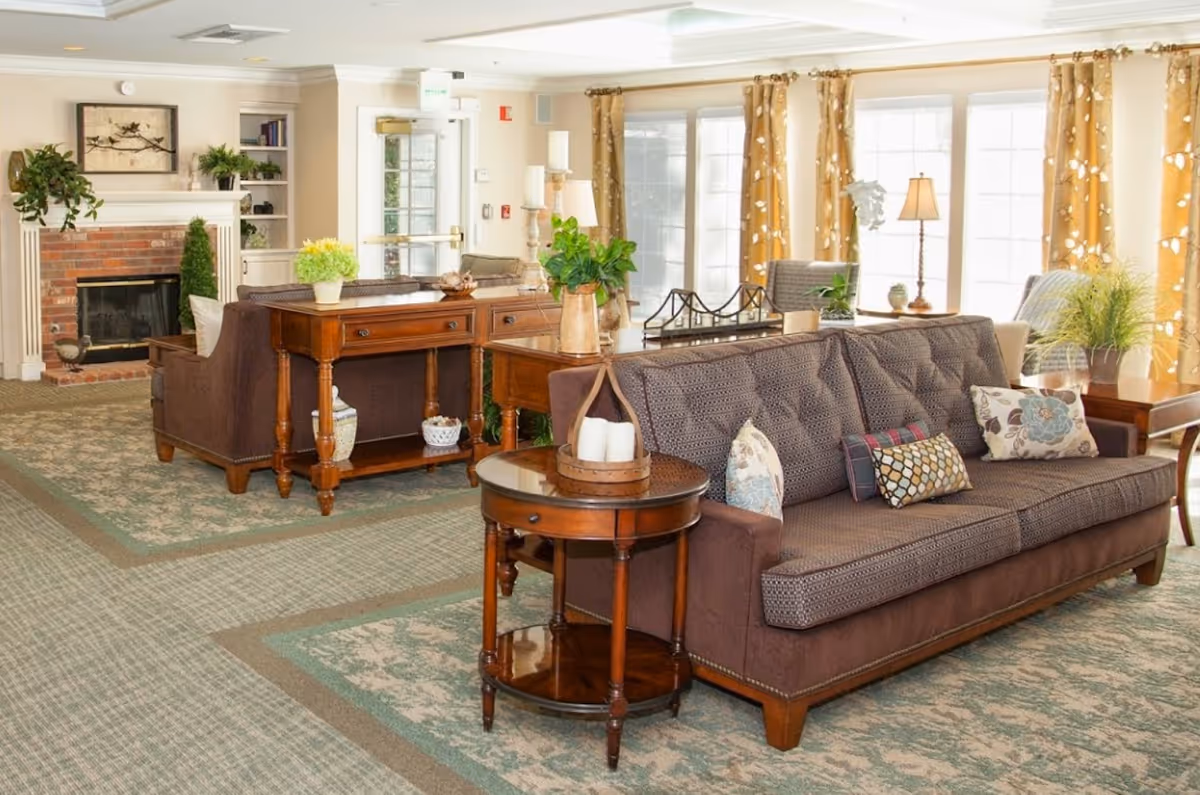 Bright communal living room with sofas, wooden side tables, plants, large windows with curtains, and a fireplace.