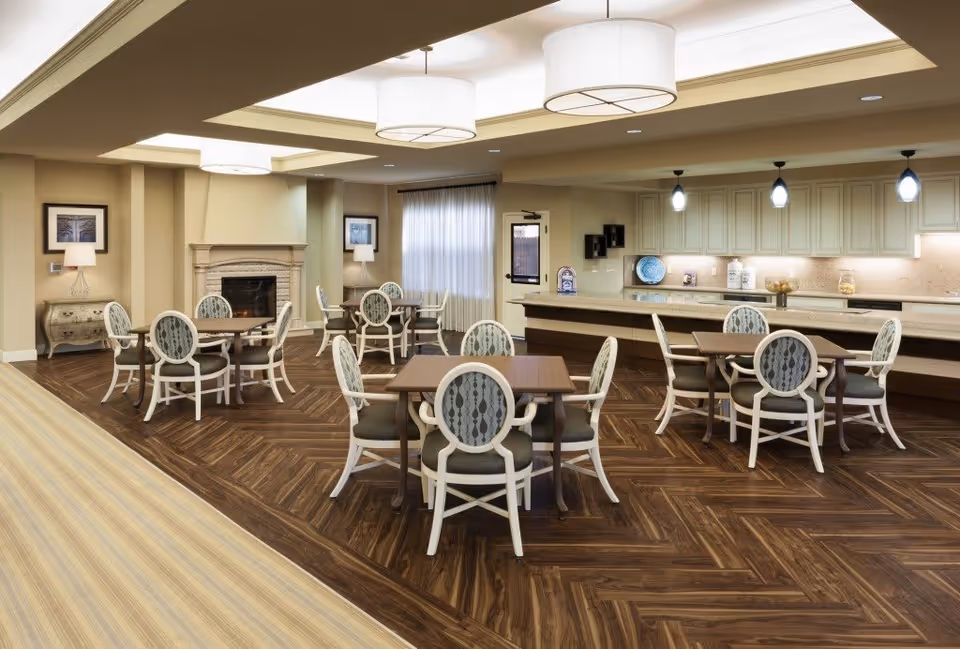 A spacious dining area with multiple square wooden tables surrounded by white chairs with patterned backs and dark cushions. The room features a fireplace with a mantle, framed artwork on the walls, and a large window with sheer curtains. The floor has a dark wood herringbone pattern, and the ceiling has recessed lighting and large round pendant lights. A long counter with cabinets and decorative items is visible in the background.