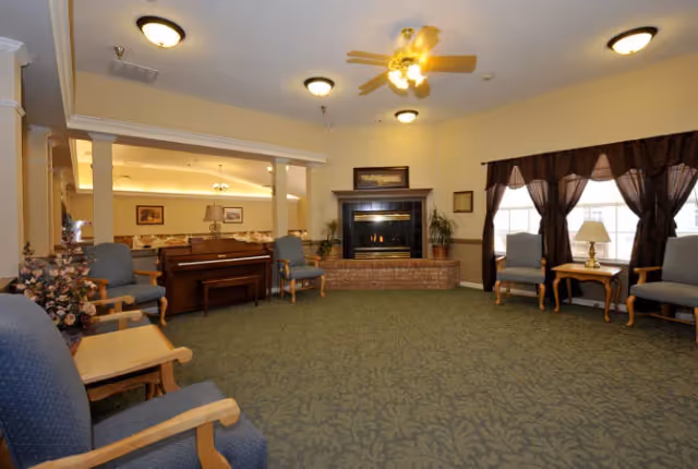 A spacious living room area with green patterned carpet, several blue upholstered chairs with wooden armrests arranged around the room, a brick fireplace with a fire burning, a piano, and large windows with dark curtains letting in natural light. The ceiling has multiple light fixtures and a ceiling fan.