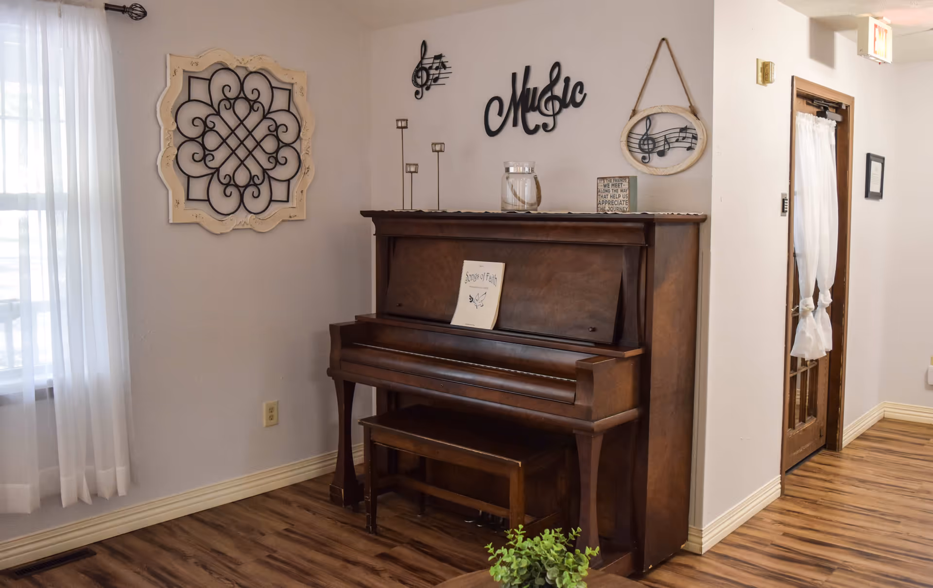 A cozy corner of a room featuring a wooden upright piano with a matching bench. On top of the piano are decorative items including candle holders, a glass jar, and a small sign. Above the piano, wall decorations include musical notes, the word 'Music' in cursive, and a framed musical note design. To the left, a window with sheer white curtains lets in natural light. The floor is wood, and a wooden door with a white curtain is visible to the right.