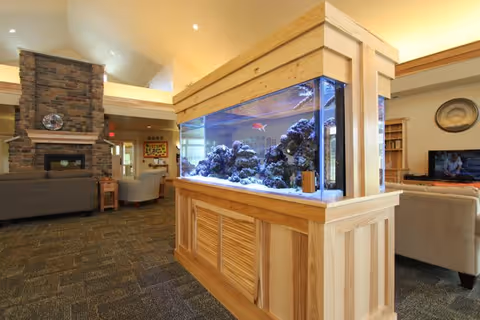 A spacious living area in an assisted living facility featuring a large built-in aquarium with colorful fish and coral. The room has a stone fireplace, comfortable seating including sofas and armchairs, carpeted flooring, and warm lighting. There is a television on the right side and decorative elements on the walls.