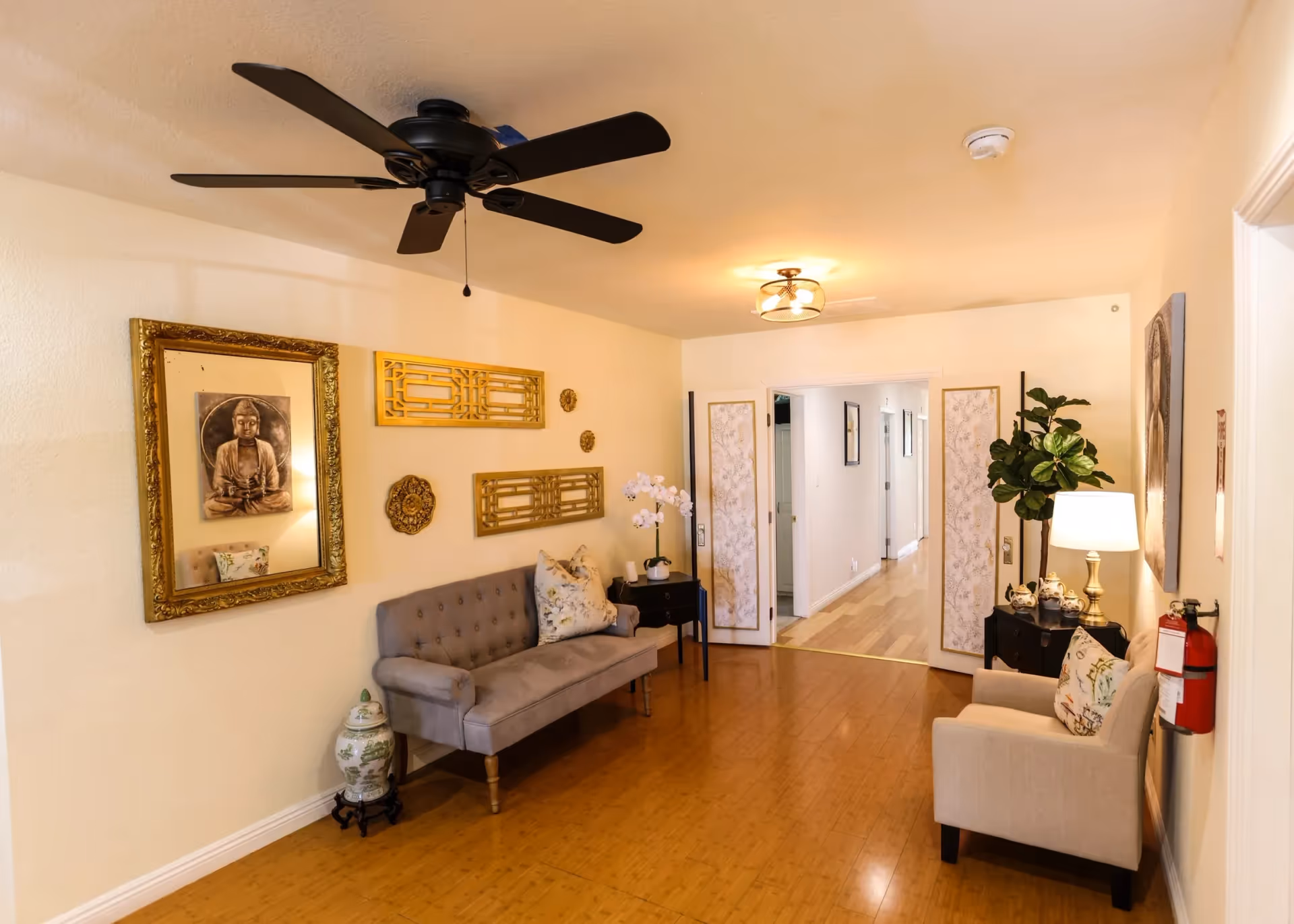 A cozy interior room with wooden flooring, featuring a gray tufted sofa with floral pillows on the left wall, decorative golden wall art, a large framed picture of a Buddha, and a ceiling fan above. On the right side, there is a beige armchair with a floral pillow, a black side table with a lamp and decorative items, a potted plant, and a fire extinguisher mounted on the wall. The room opens into a hallway with wooden floors and white walls.