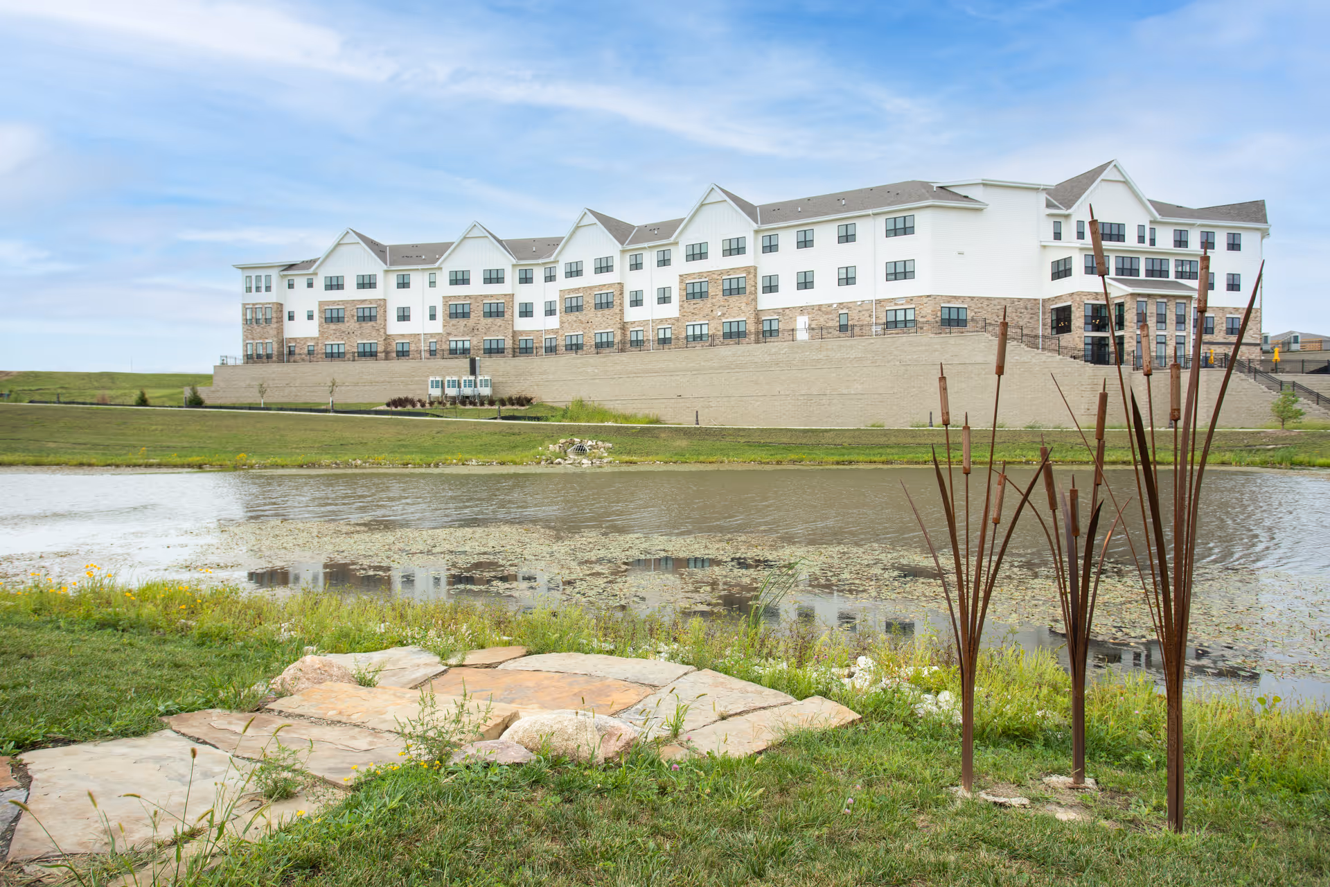 A large, multi-story senior living facility building with white and brick exterior walls situated on a grassy hill overlooking a pond with aquatic plants. In the foreground, there is a stone pathway and tall metal sculptures resembling cattails. The sky is partly cloudy.