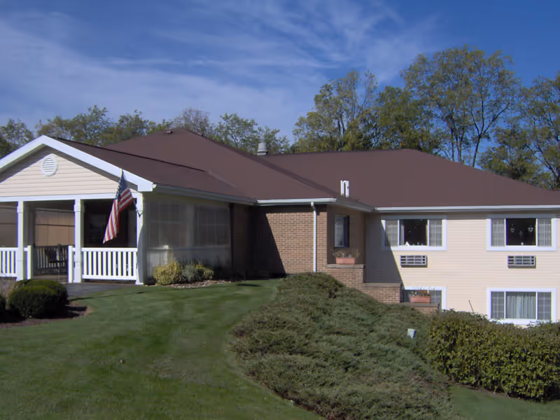 Exterior view of a single-story residential building with beige siding and a brown roof, featuring a covered porch with white railings and an American flag. The building is surrounded by green grass, shrubs, and trees under a blue sky with some clouds.