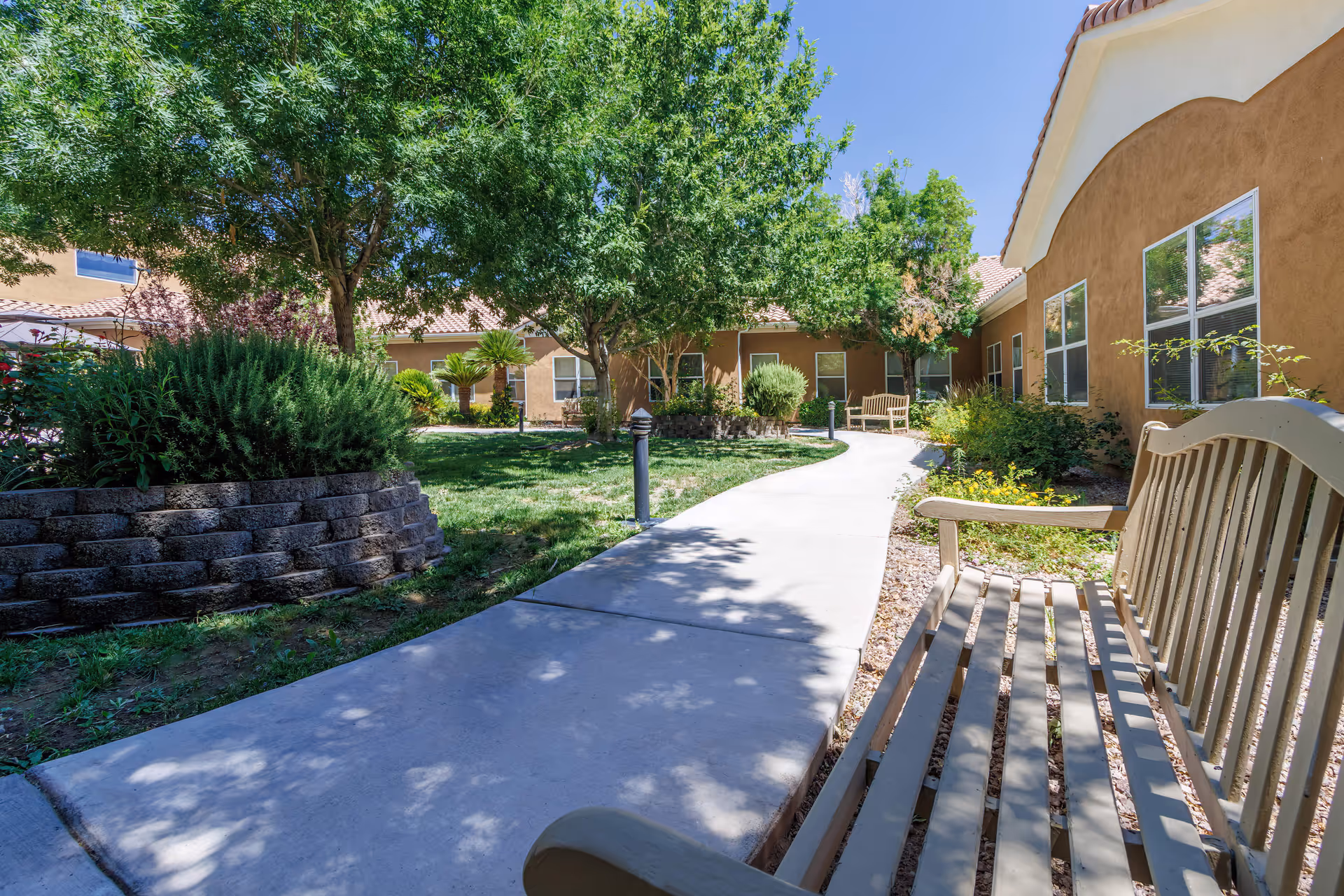 A sunny outdoor courtyard area with a concrete pathway winding through green grass and landscaped plants. There are several trees providing shade and benches along the path. The surrounding buildings have tan stucco walls and tiled roofs under a clear blue sky.