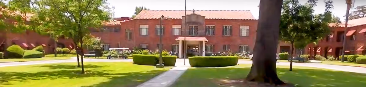 Front exterior view of Claremont Manor Retirement Community showing a large brick building with a tiled roof, surrounded by green lawns, trees, and bushes under a clear sky.