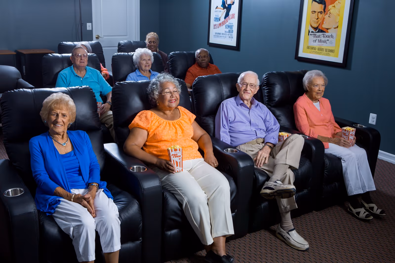A group of elderly people sitting in black leather recliners in a small movie theater room. Some are holding popcorn containers. The walls are painted dark teal and have framed vintage movie posters. The carpet is brown with a subtle pattern.
