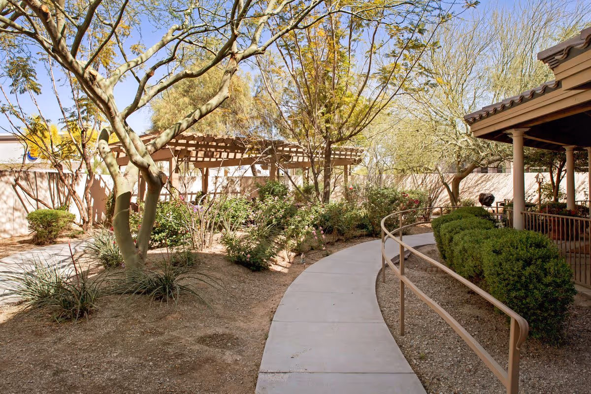 Curved concrete walkway with a handrail through a landscaped courtyard featuring trees, shrubs and a wooden pergola.