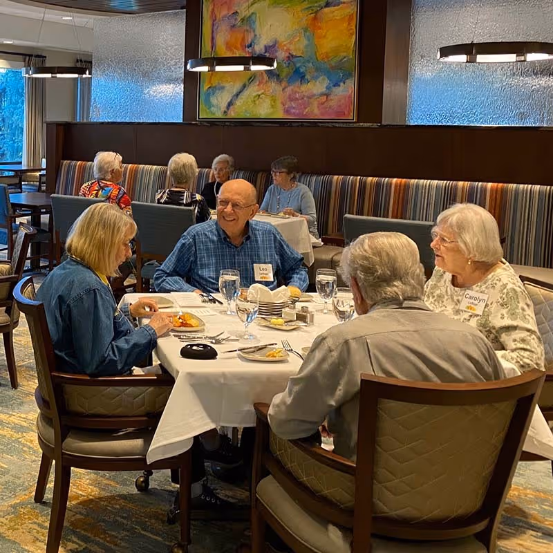 A group of elderly people sitting around a table in a dining room at Frasier Retirement Community, enjoying a meal and conversation. The table is set with plates, glasses, and silverware. The room has striped cushioned seating along the wall, modern hanging lights, and colorful abstract artwork on the wall.
