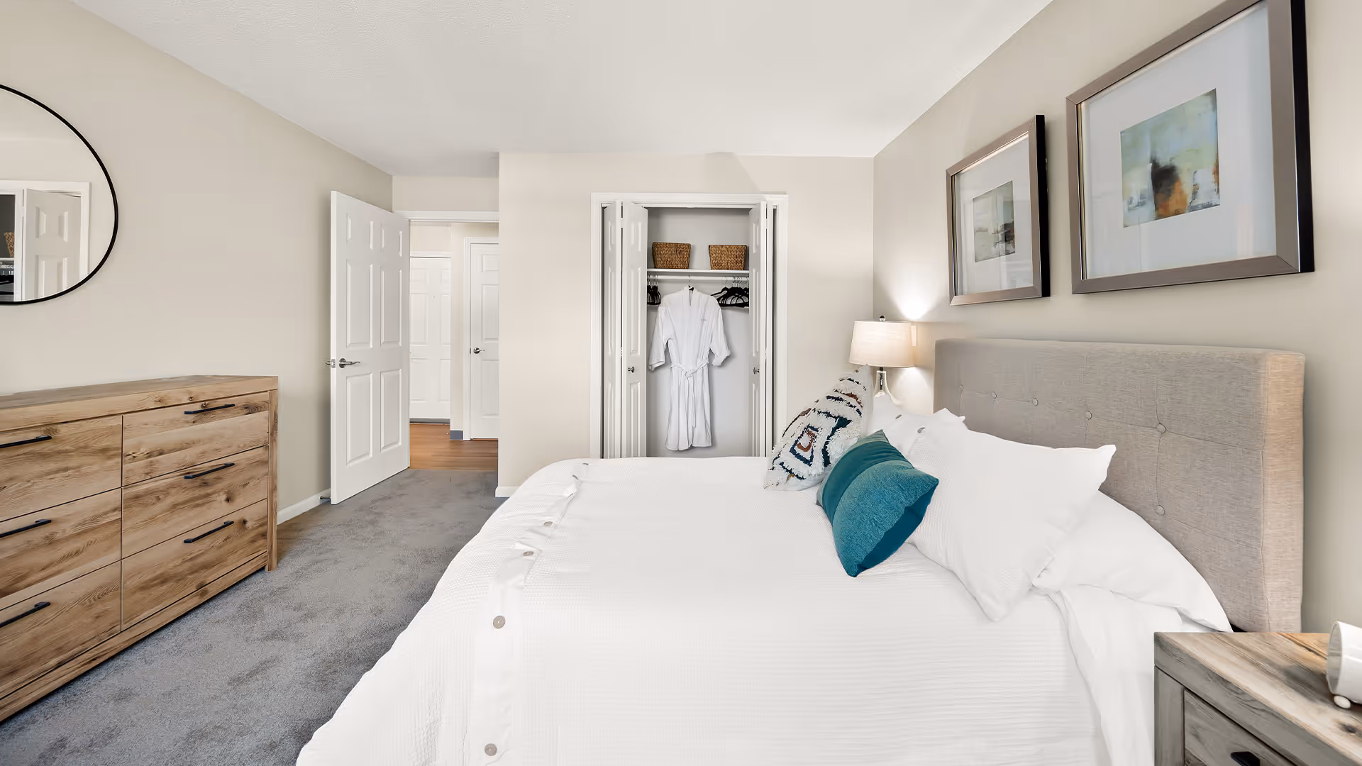 A neatly arranged bedroom with a large bed featuring white bedding and teal accent pillows. There is a wooden dresser with black handles on the left side and a round mirror above it. The closet doors are open, revealing a white robe hanging inside. Two framed abstract artworks hang above the bed, and a bedside table with a lamp is on the right side. The room has beige walls and gray carpet flooring.