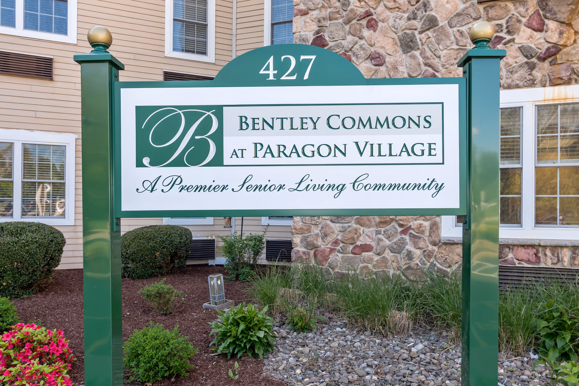 Green and white sign for Bentley Commons at Paragon Village, a premier senior living community, mounted on green posts with a building and landscaping in the background.