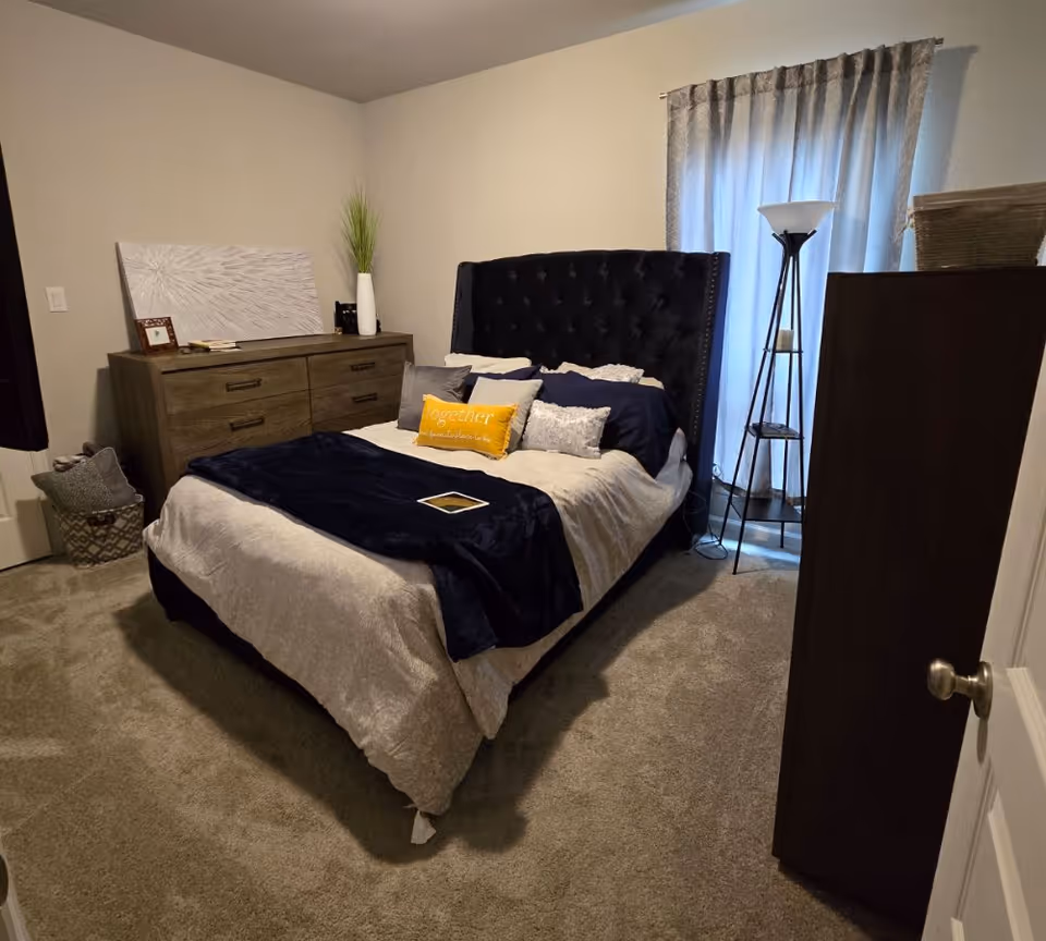 A cozy bedroom with a large bed featuring a dark tufted headboard, beige and navy bedding, and multiple pillows including a yellow one with the word 'Together' on it. There is a wooden dresser with decorative items and a tall floor lamp near a window with curtains. The room has beige carpet and neutral-colored walls.
