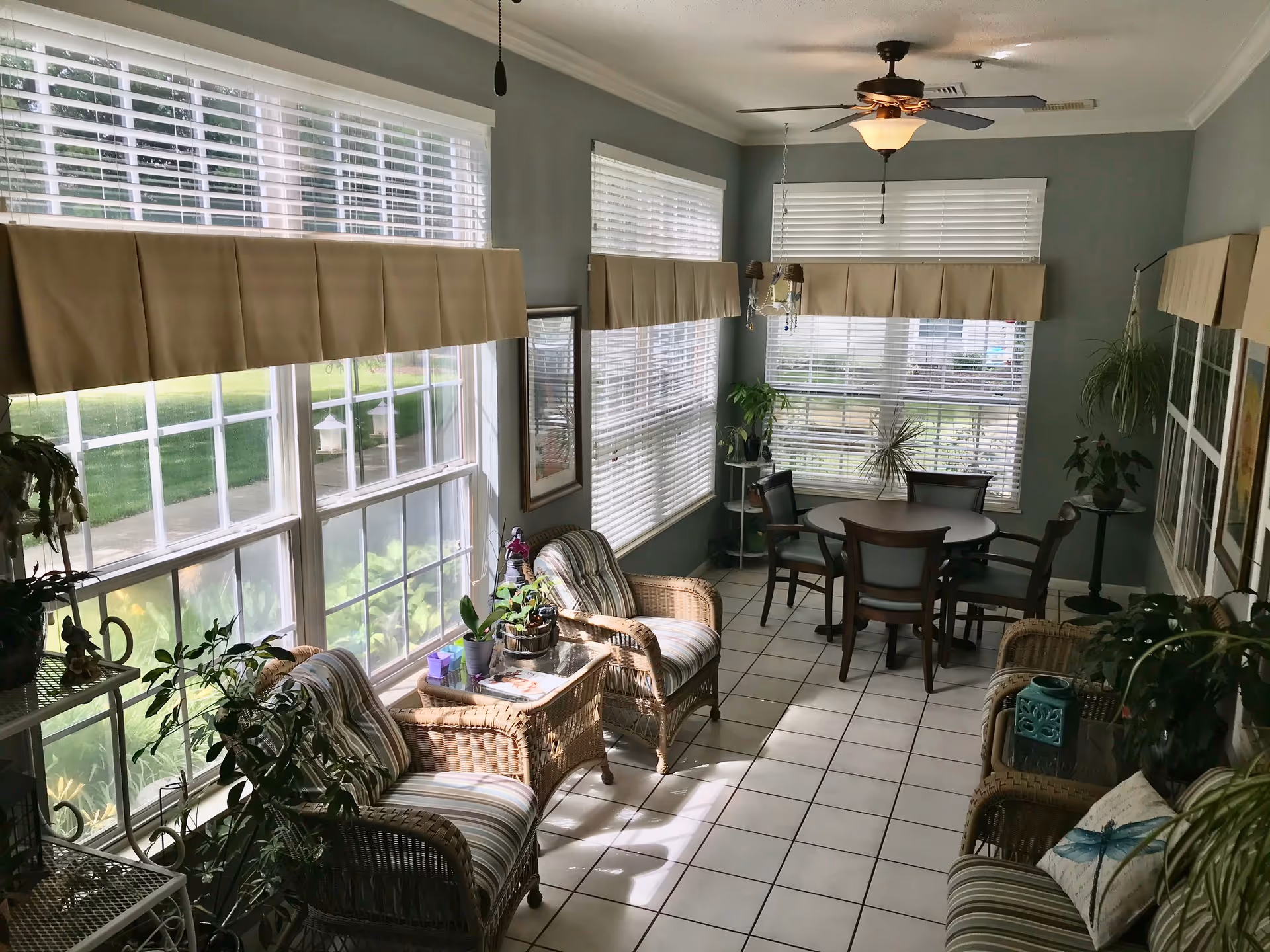 A bright sunroom with large windows covered by white blinds and beige valances. The room features wicker chairs with striped cushions, a small round table with four chairs, several potted plants, and a ceiling fan with a light fixture. The floor is tiled with white square tiles.