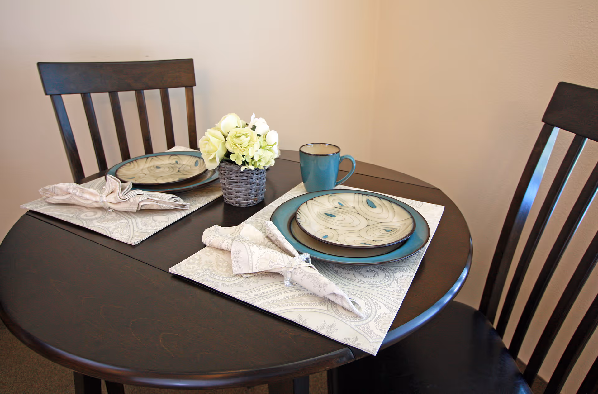 Small round dining table set for two with plates, cloth napkins, a mug, and a small flower centerpiece.