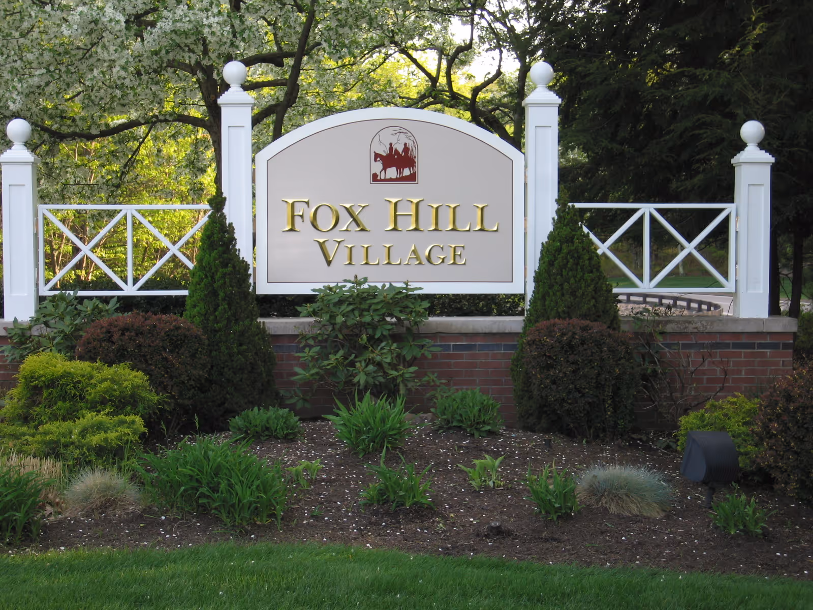 A landscaped entrance sign for Fox Hill Village featuring white posts with decorative finials, a white fence with crossbars, and a sign with gold lettering. The area around the sign is planted with various shrubs and greenery, with trees in the background.