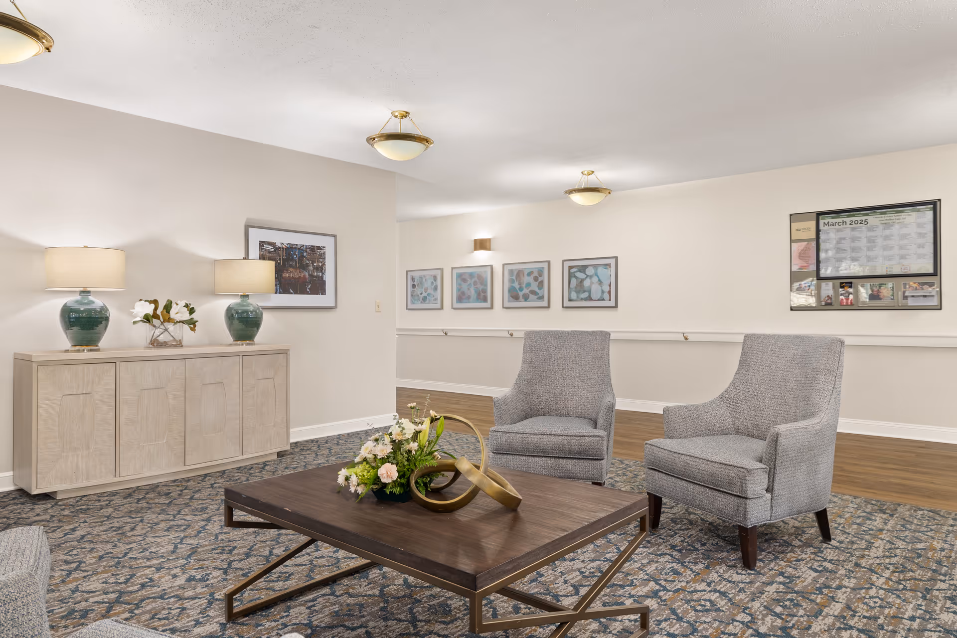 A senior living facility common area with two gray upholstered armchairs and a wooden coffee table with a floral arrangement and decorative sculpture. The room has a patterned carpet, light beige walls adorned with framed artwork, a sideboard with two green lamps and a flower arrangement, and ceiling light fixtures. A bulletin board with a calendar and photos is visible on the far wall.