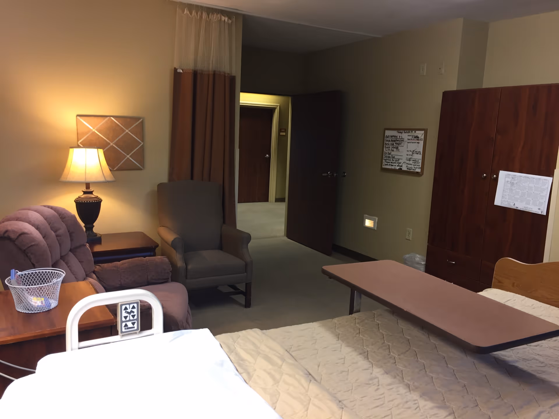A nursing facility room with a hospital bed covered in a beige quilt, an overbed table, a brown armchair, a purple recliner, a wooden nightstand with a lamp, a wooden wardrobe, and an open door leading to a hallway.