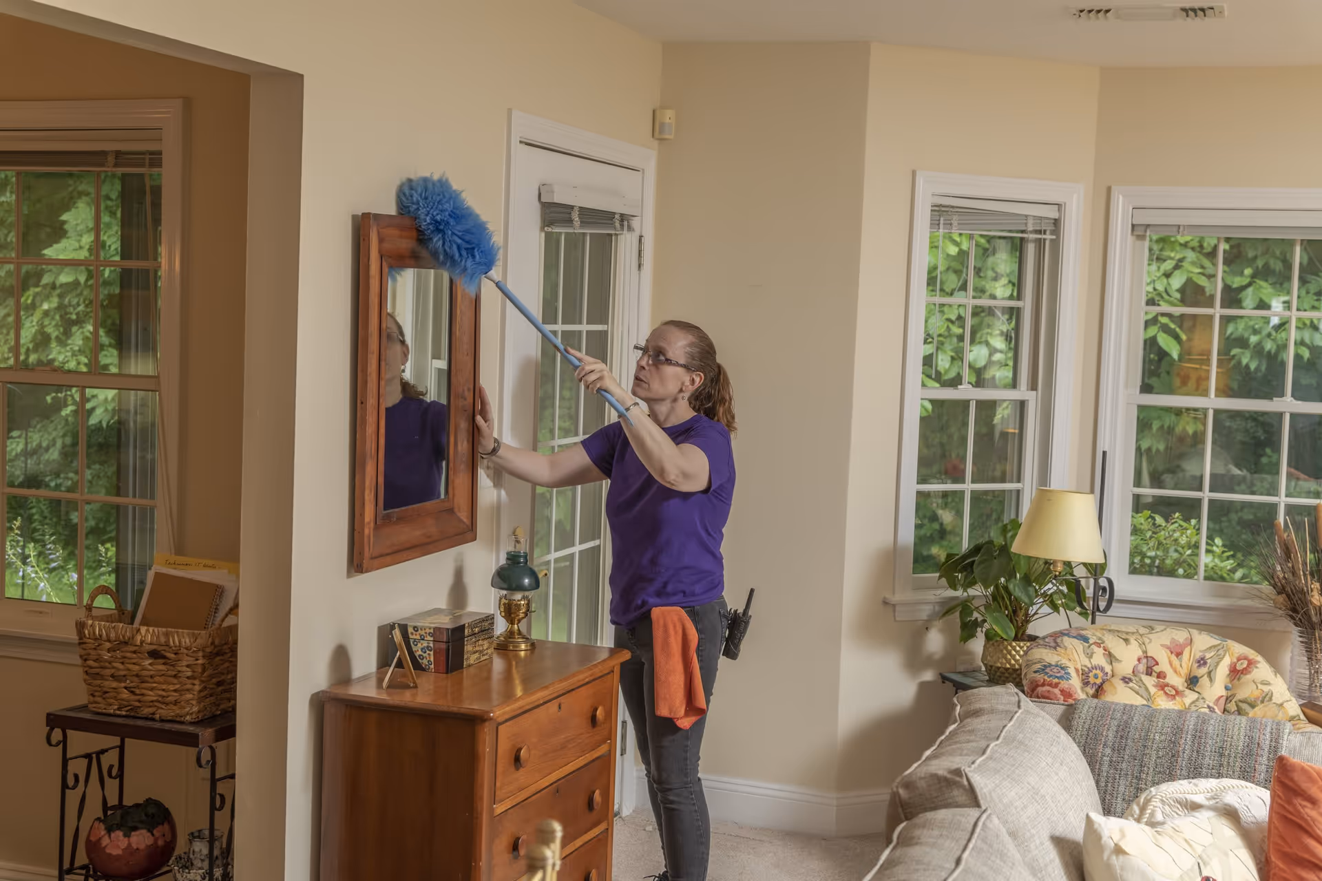 A woman wearing a purple shirt and glasses dusts a wooden framed mirror on a beige wall in a cozy living room. The room features large windows with white frames showing green foliage outside, a wooden chest of drawers with decorative items, a floral armchair, a table lamp, and a beige sofa with cushions.