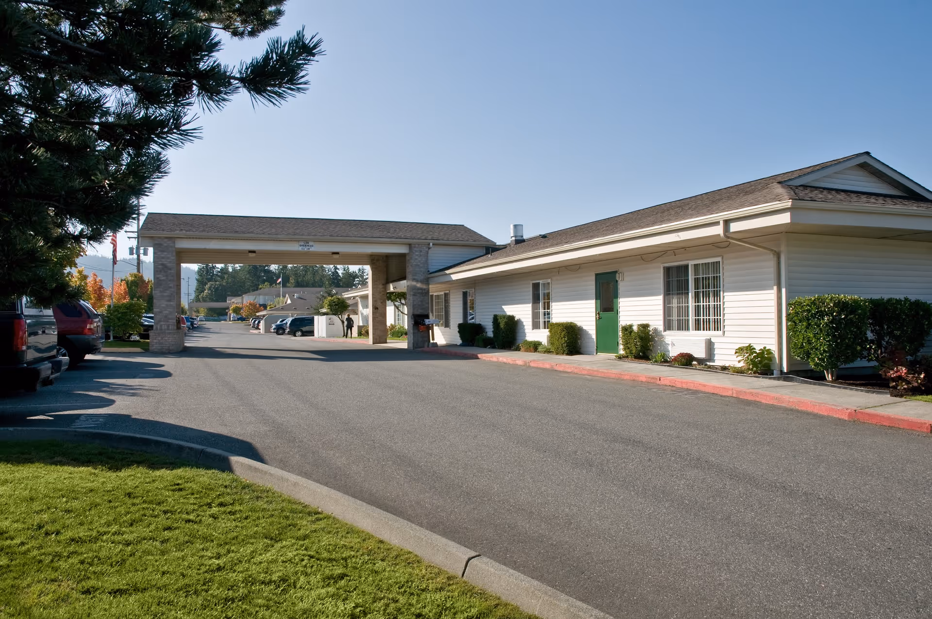 Exterior view of a single-story building with white siding and a green door, part of a senior living facility. There is a covered entrance with a driveway and several parked cars along the side. Trees and bushes are visible around the building under a clear blue sky.