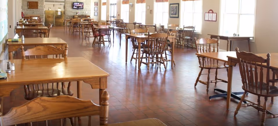Spacious dining room with multiple wooden tables and chairs arranged on a tiled floor and windows along the right wall.