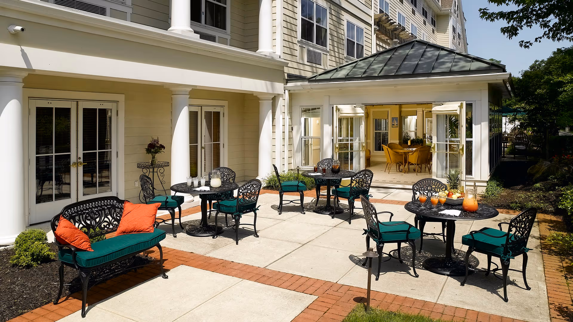 Outdoor patio area at a senior living facility with black wrought iron tables and chairs featuring green cushions and orange pillows. The patio is paved with concrete and bordered by red bricks. There are drinks and fruit on the tables, and the patio is adjacent to a building with large windows and glass doors leading inside.