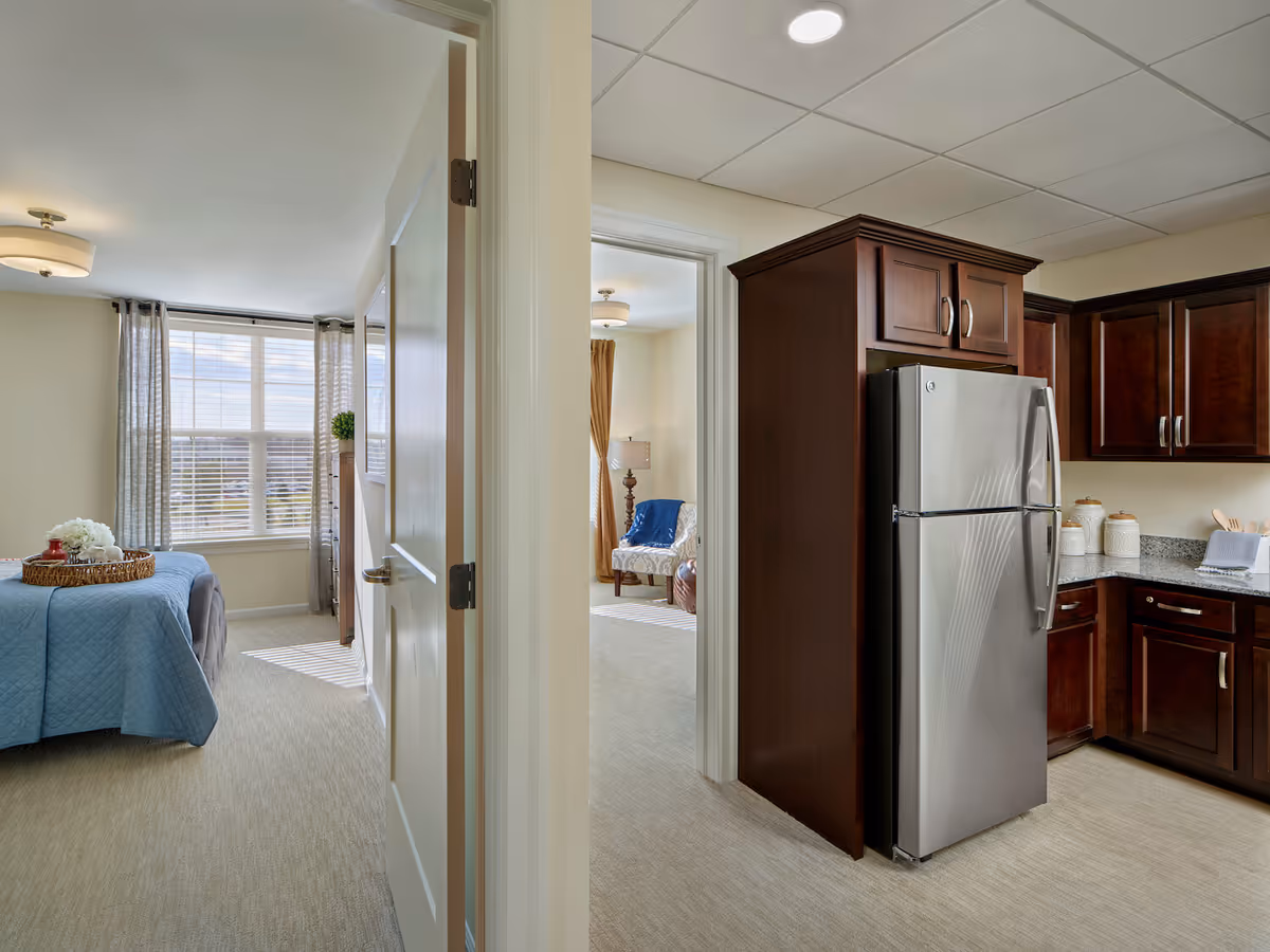 View of an interior space showing a kitchen with dark wood cabinets and a stainless steel refrigerator on the right, and two adjacent rooms visible through open doors. One room has a bed with a blue quilt and a window with sheer curtains, while the other room has a chair with a blue throw and a floor lamp.