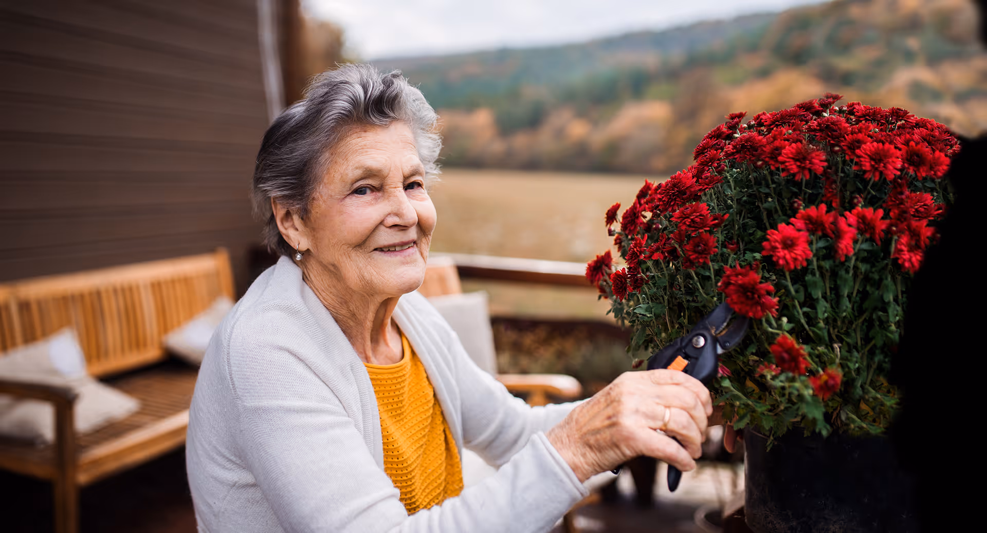 An elderly woman with gray hair wearing a yellow top and light gray cardigan is smiling while trimming red flowers with garden shears on a wooden patio. There is a wooden bench with cushions in the background and a scenic view of trees and hills.