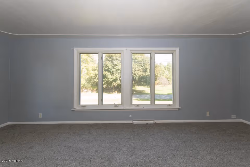 Empty room with gray carpet and light blue walls featuring a large window with four panes showing green trees and a lawn outside.