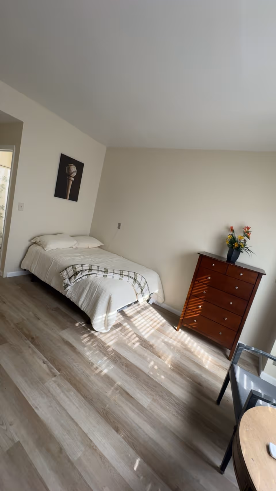 Bright single bedroom with a bed against a pale wall, a wooden chest of drawers with flowers, and sunlight on the wood floor.