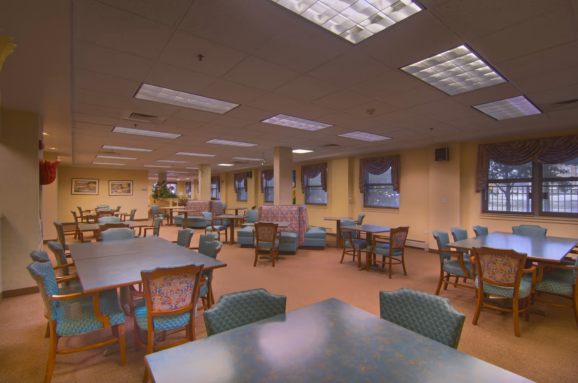 A spacious dining room in a senior living facility with multiple tables and chairs arranged neatly. The room has beige walls, carpeted floors, and several windows with valance curtains allowing natural light. The ceiling has fluorescent lighting panels, and there are some decorative plants and framed pictures on the walls.