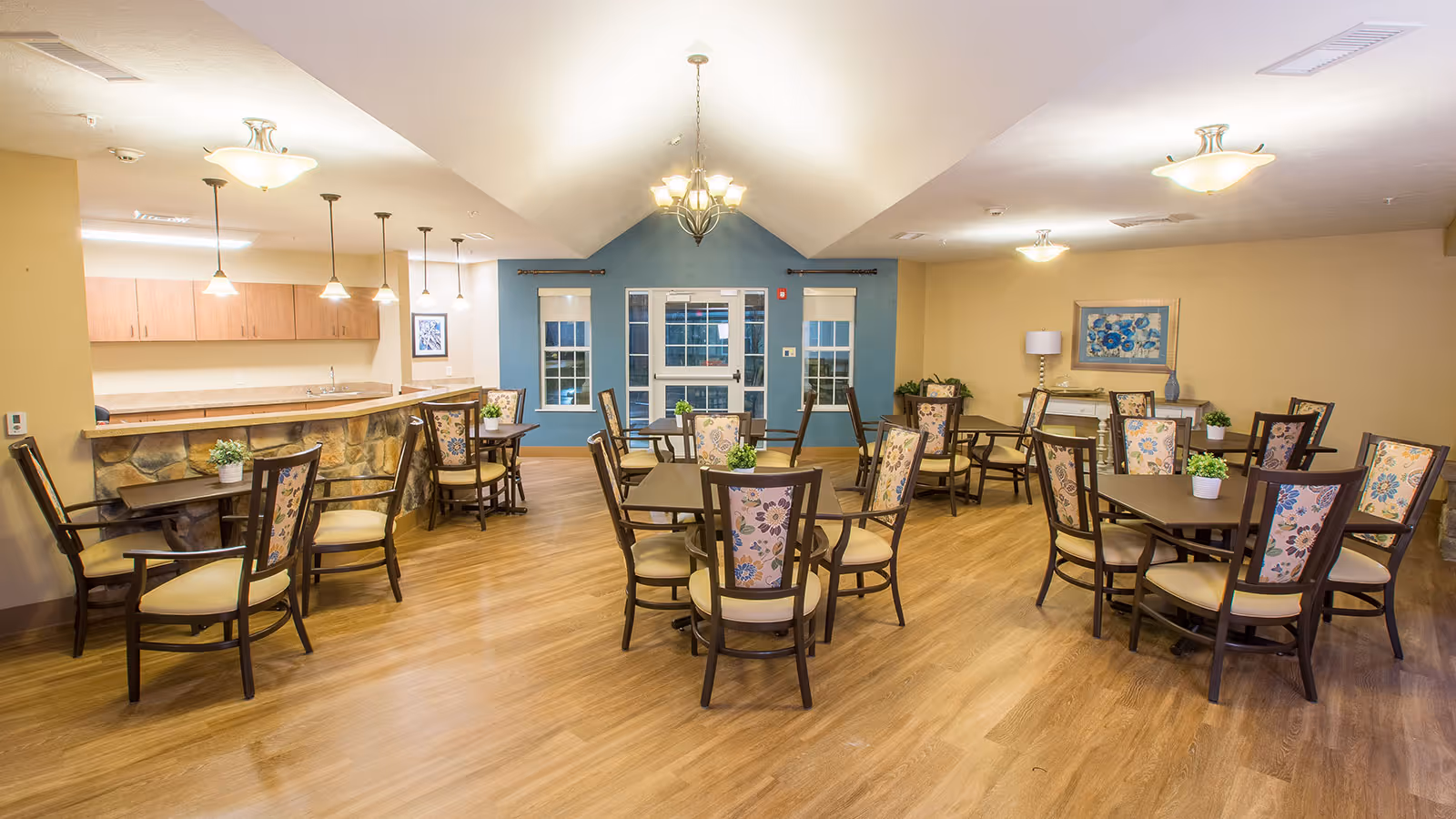 A spacious dining room with multiple square tables surrounded by wooden chairs with floral upholstered backs and beige cushions. The room features wood flooring, a stone-faced counter with a kitchen area behind it, and a blue accent wall with windows and a door. The ceiling has several light fixtures including chandeliers and pendant lights. There are framed artworks and potted plants decorating the space.