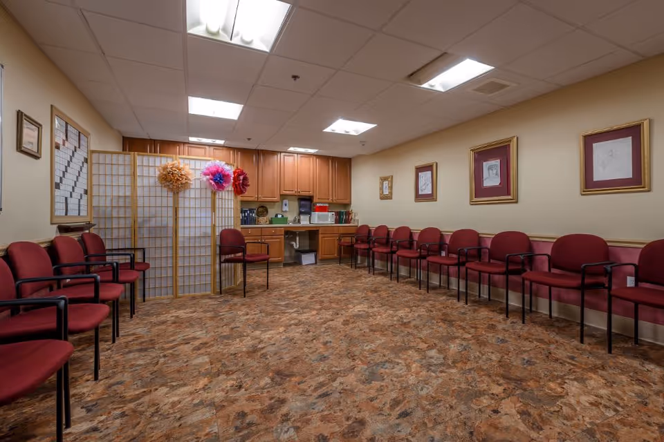 A waiting room with maroon chairs lined up along two walls, a wooden folding screen decorated with large paper flowers, wooden cabinets with a countertop holding a microwave and other items, and framed artwork on the walls.
