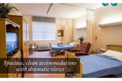A spacious and clean nursing center room with a bed, a reclining chair, a small table with chairs, a television on a wooden stand, and a large window with curtains allowing natural light to enter.