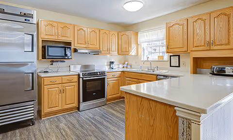 A bright kitchen with wooden cabinets, a stainless steel refrigerator, a microwave, an electric stove, a coffee maker, a toaster, and a large countertop island. The floor has wood-like vinyl flooring, and there is a window above the sink letting in natural light.