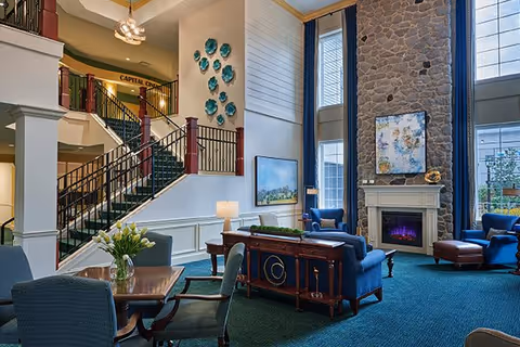 Spacious communal lounge with upholstered chairs and tables, a stone fireplace beneath tall windows, and a staircase leading to an upper level.