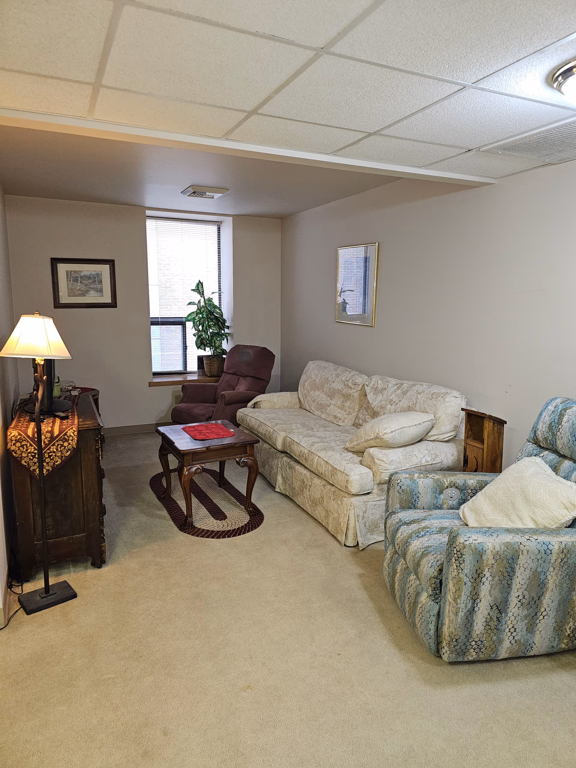A cozy living room with a beige carpet, a cream-colored sofa with cushions, a blue patterned recliner chair, and a maroon recliner near a window with blinds. There is a wooden coffee table with a red cloth on top, a floor lamp with a beige shade, a wooden cabinet with a decorative cloth, a potted plant on the windowsill, and framed pictures on the walls.