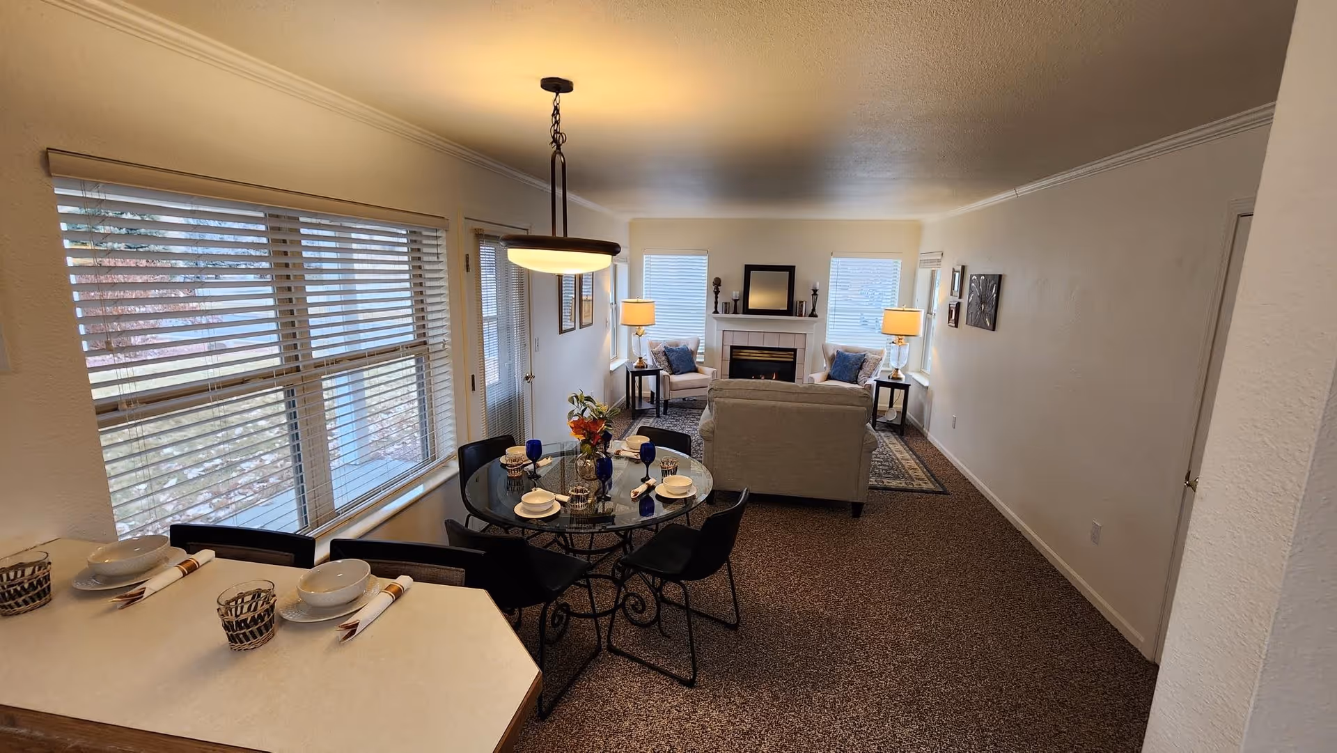Interior view of a senior living facility showing a combined dining and living area. The dining area features a round glass table set with plates, bowls, napkins, and blue glasses, surrounded by black chairs. Adjacent to it is a kitchen counter with place settings. The living area has a beige sofa facing a fireplace with a mirror above it, flanked by two armchairs and side tables with lamps. Large windows with blinds allow natural light into the room.