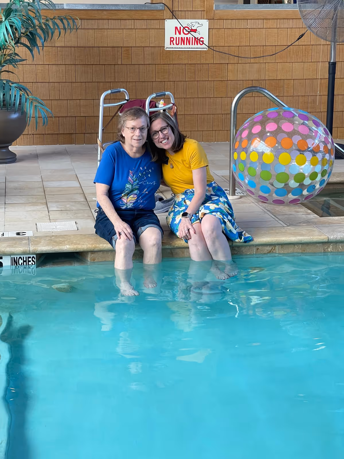 Two women sitting at the edge of an indoor swimming pool with their feet in the water. One woman is wearing a blue shirt and black shorts, and the other is wearing a yellow shirt and a patterned skirt. Behind them is a walker, a large potted plant, a colorful polka-dot beach ball, and a sign on the wall that says 'NO RUNNING'.