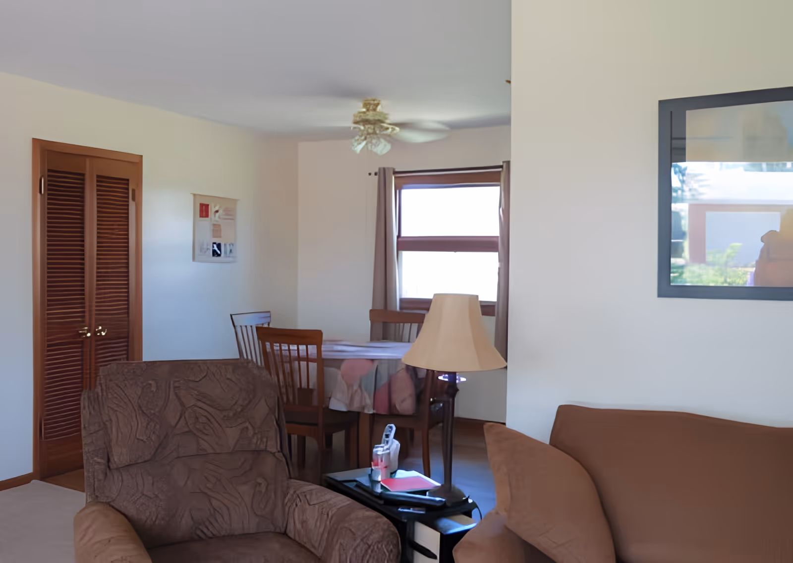 A cozy living room area with a brown armchair and a brown sofa. A small side table with a lamp, a remote control, and some papers is placed between the armchair and sofa. In the background, there is a dining area with a wooden table covered with a tablecloth and four wooden chairs. A window with curtains and a ceiling fan are also visible. The walls are light-colored, and there is a framed picture on the wall.