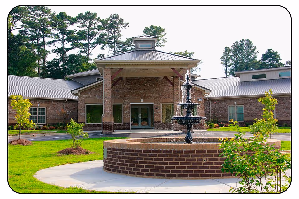 Front exterior view of Thomas Kelly Senior Living Community building with a brick facade, metal roof, and a multi-tiered water fountain in the foreground surrounded by green grass and small trees.