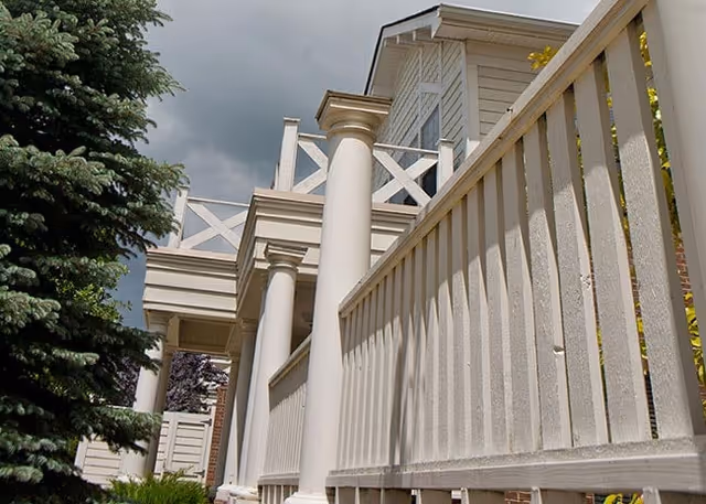 Exterior view of a light-colored building with white columns, a balustrade railing, and an evergreen tree under a cloudy sky.