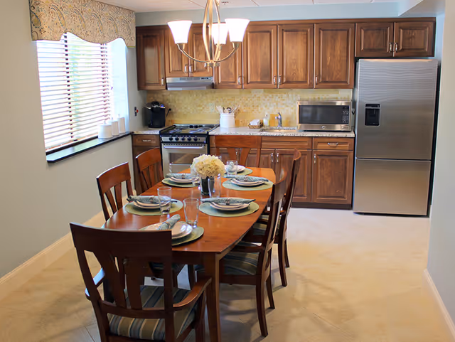 A kitchen and dining area with wooden cabinets, a stainless steel refrigerator, stove, microwave, and a wooden dining table set for six with plates, glasses, and a flower centerpiece. A window with blinds and a valance is on the left wall.